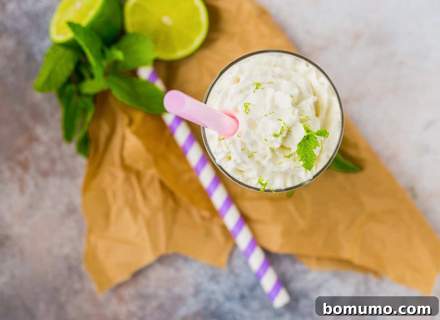Mojito Dream Shake 3 A glass of whipped cream-topped mojito milkshake adorned with lime zest and mint, a pink striped straw, two green striped straws, vibrant lime slices, and fresh mint leaves artfully arranged on brown paper, suggesting a refreshing summer vibe.