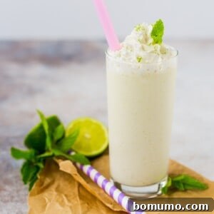 A tall glass of mojito milkshake topped with whipped cream and a mint leaf, with a pink straw. Beside it are a lime half, mint sprigs, and a purple-striped straw.