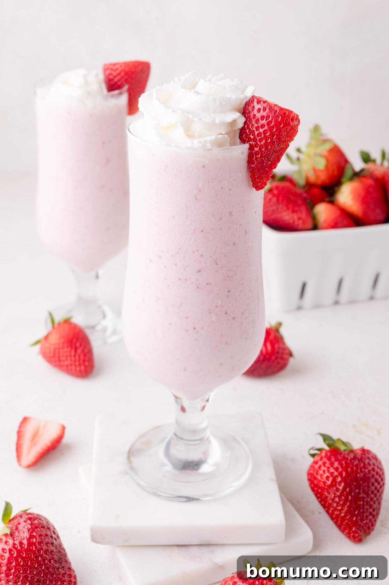 Velvet Strawberry Indulgence 2 A tall glass of strawberry milkshake topped with a swirl of fresh whipped cream and a perfect strawberry slice. Scattered around are fresh, vibrant strawberries, and another glass of milkshake subtly blurred in the background, creating a refreshing summer scene.