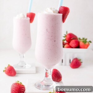 Two enticing glasses of strawberry milkshake, perfectly topped with swirls of whipped cream and garnished with fresh strawberries. Whole strawberries are scattered artfully around, with a container of fresh berries blurred in the background, completing a delicious and refreshing scene.