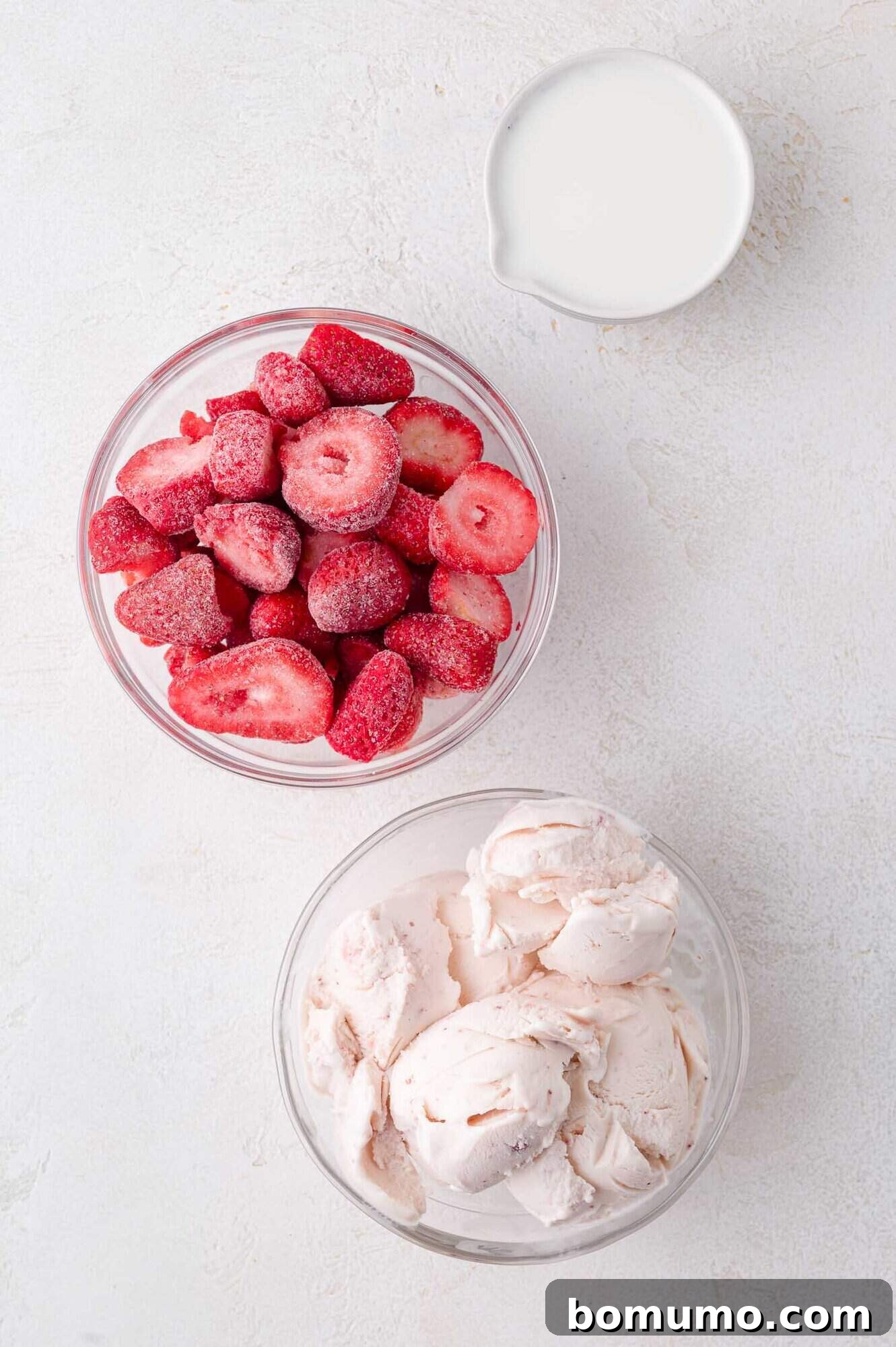 Velvet Strawberry Indulgence 3 A vibrant display of fresh ingredients for a homemade strawberry milkshake, featuring a bowl of perfectly frozen strawberries, a container of rich strawberry ice cream, and a small pitcher of fresh milk, all arranged appealingly on a clean white surface.