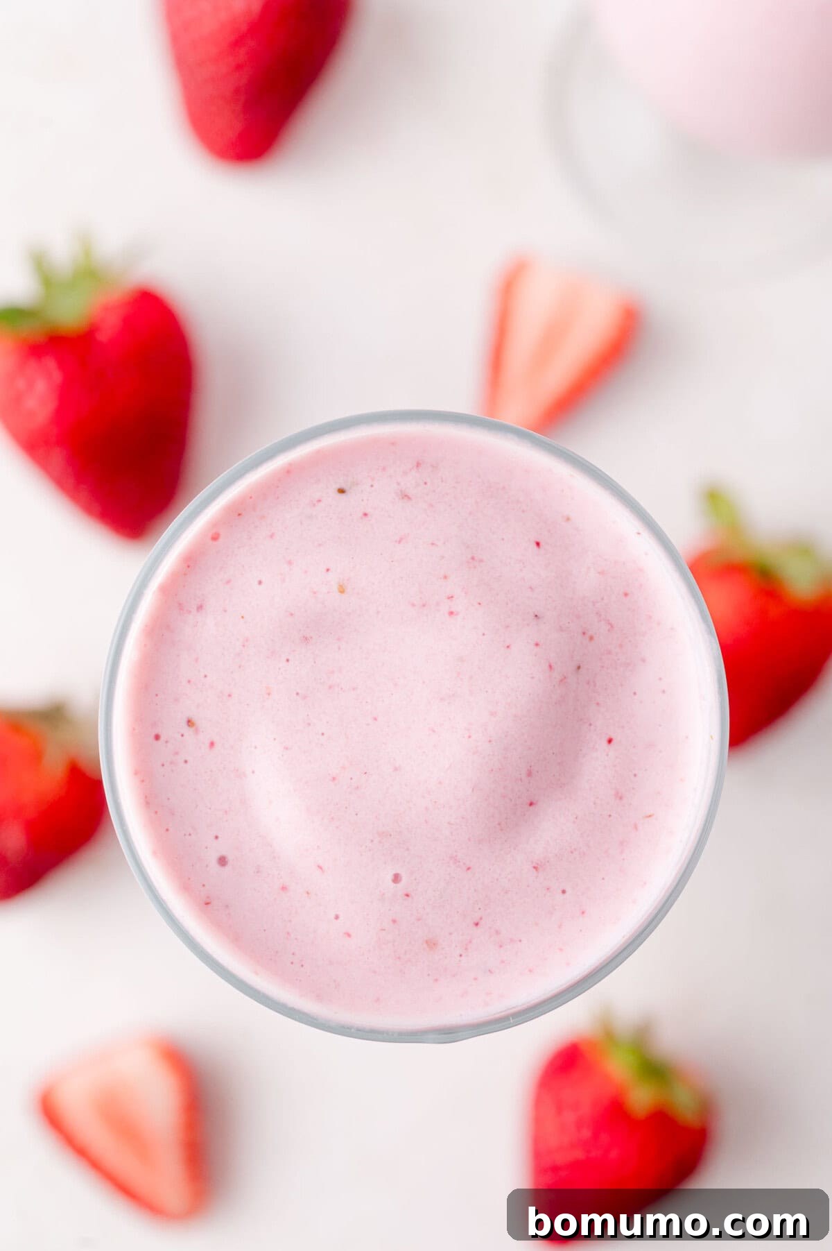 Velvet Strawberry Indulgence 7 A beautifully presented glass filled with a pink strawberry milkshake, elegantly surrounded by a scattering of whole and halved fresh strawberries on a clean white surface, highlighting its natural goodness.