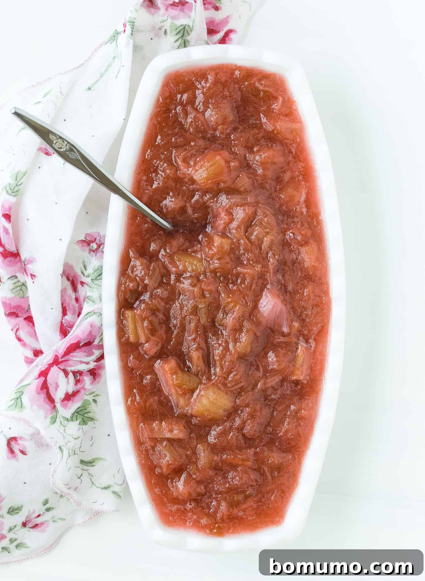 A serving of homemade rhubarb sauce in an elegant oval white bowl, accompanied by a silver spoon.