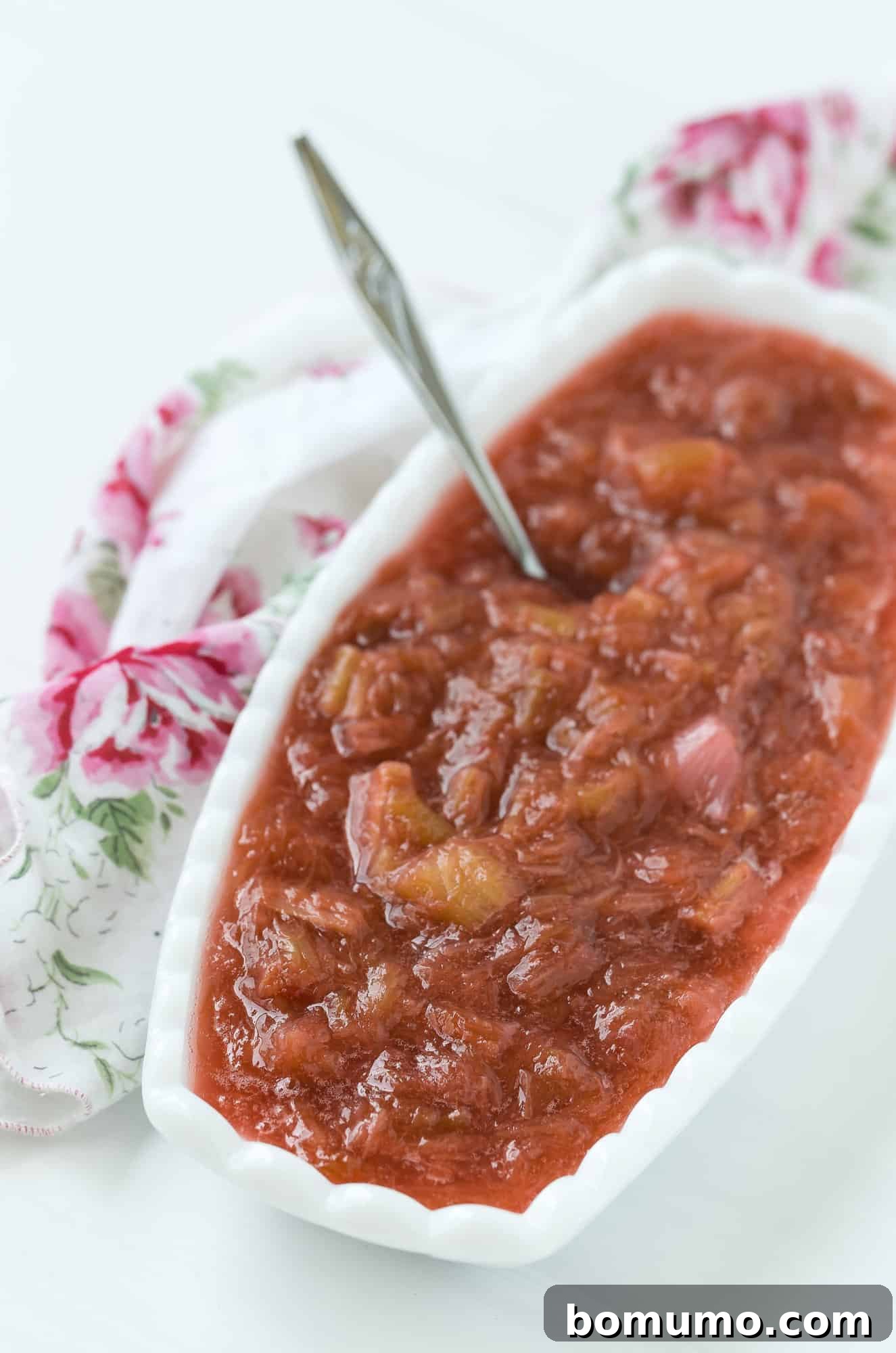 A close-up of light red homemade rhubarb sauce in an oval white bowl, with a spoon resting inside.