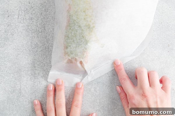 One hand holding the parchment paper packet closed, while the other hand deftly folds over and crimps the edges to create a tight, sealed package.
