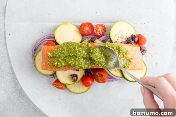 A spoon spreading rich, green pesto evenly over a salmon fillet, which rests on a bed of vibrant vegetables on a parchment paper oval.