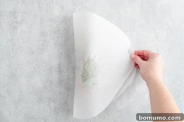 A hand carefully folds a parchment paper oval in half, enclosing the pesto salmon ingredients inside, ready for sealing.