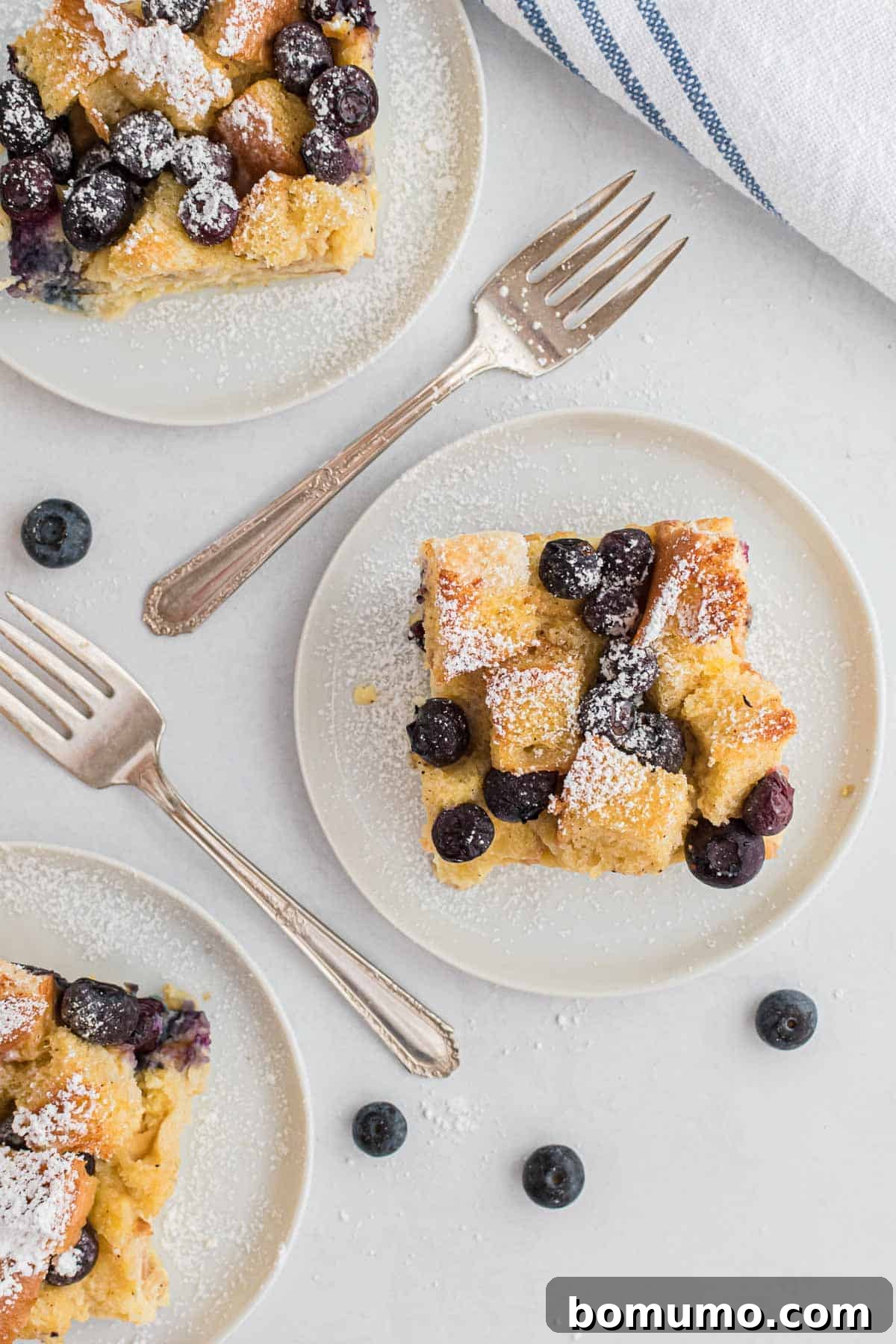 Comforting Blueberry Bread Pudding 2 Three golden-brown slices of homemade blueberry bread pudding on white plates, garnished with fresh blueberries and forks, ready to be enjoyed.