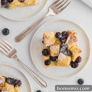 Three slices of blueberry bread pudding on round white plates, with forks and fresh blueberries nearby.