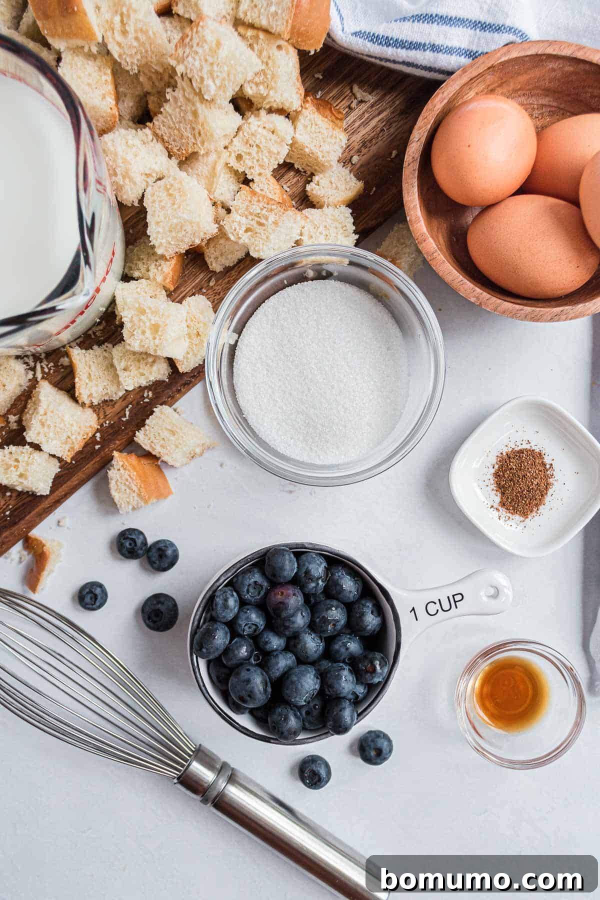 Comforting Blueberry Bread Pudding 3 A vibrant overhead shot of essential blueberry bread pudding ingredients: cinnamon sticks, neatly cubed bread, a pitcher of fresh milk, farm-fresh eggs, and plump, juicy blueberries.