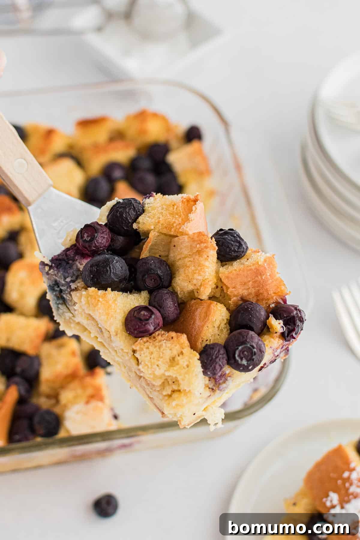 Comforting Blueberry Bread Pudding 6 A serving spoon expertly scooping a warm, custardy slice of blueberry bread pudding from a square glass baking pan, highlighting its inviting texture.