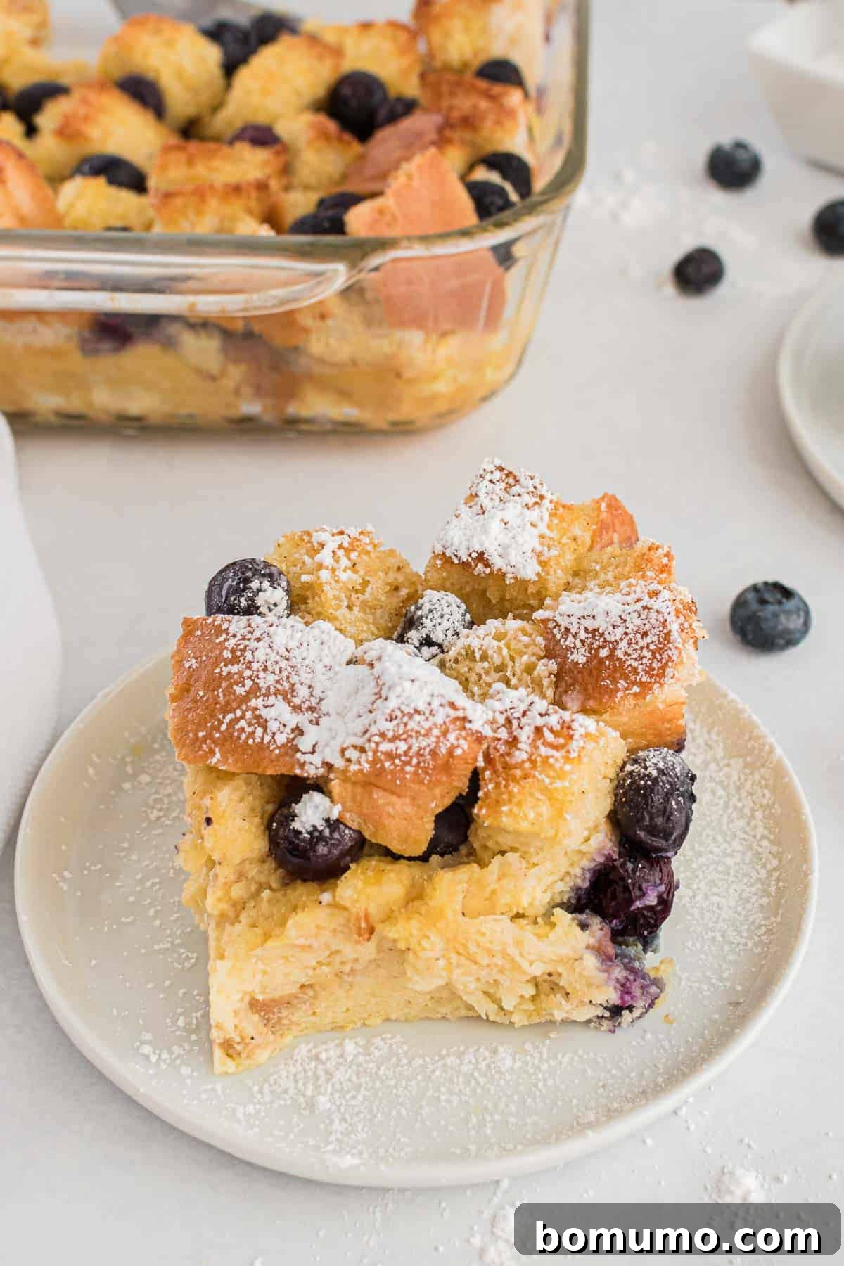 Comforting Blueberry Bread Pudding 7 A single slice of blueberry bread pudding, lightly dusted with powdered sugar, resting on a round white plate, ready for a delicious bite.