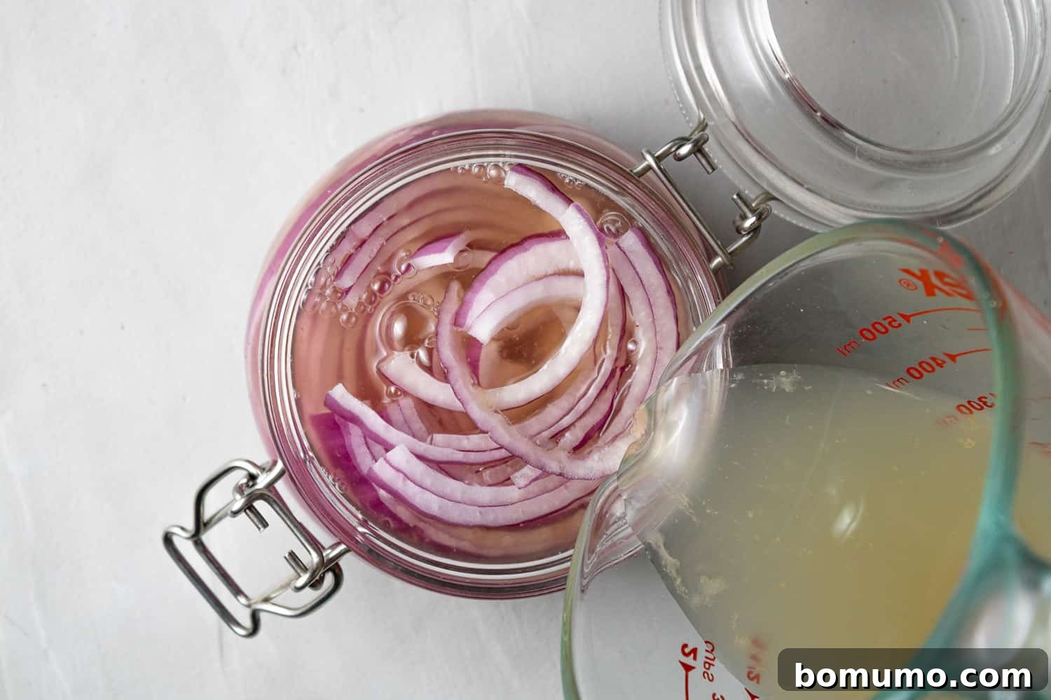 Pickling liquid being poured from a measuring cup over thinly sliced red onions packed into a glass jar.