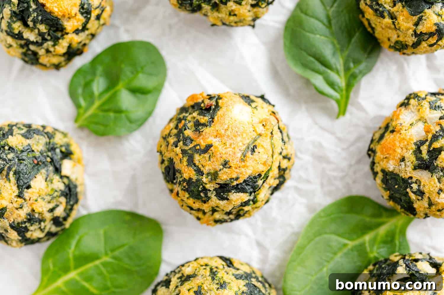 Overhead view of golden baked spinach balls surrounded by spinach leaves on a white surface.