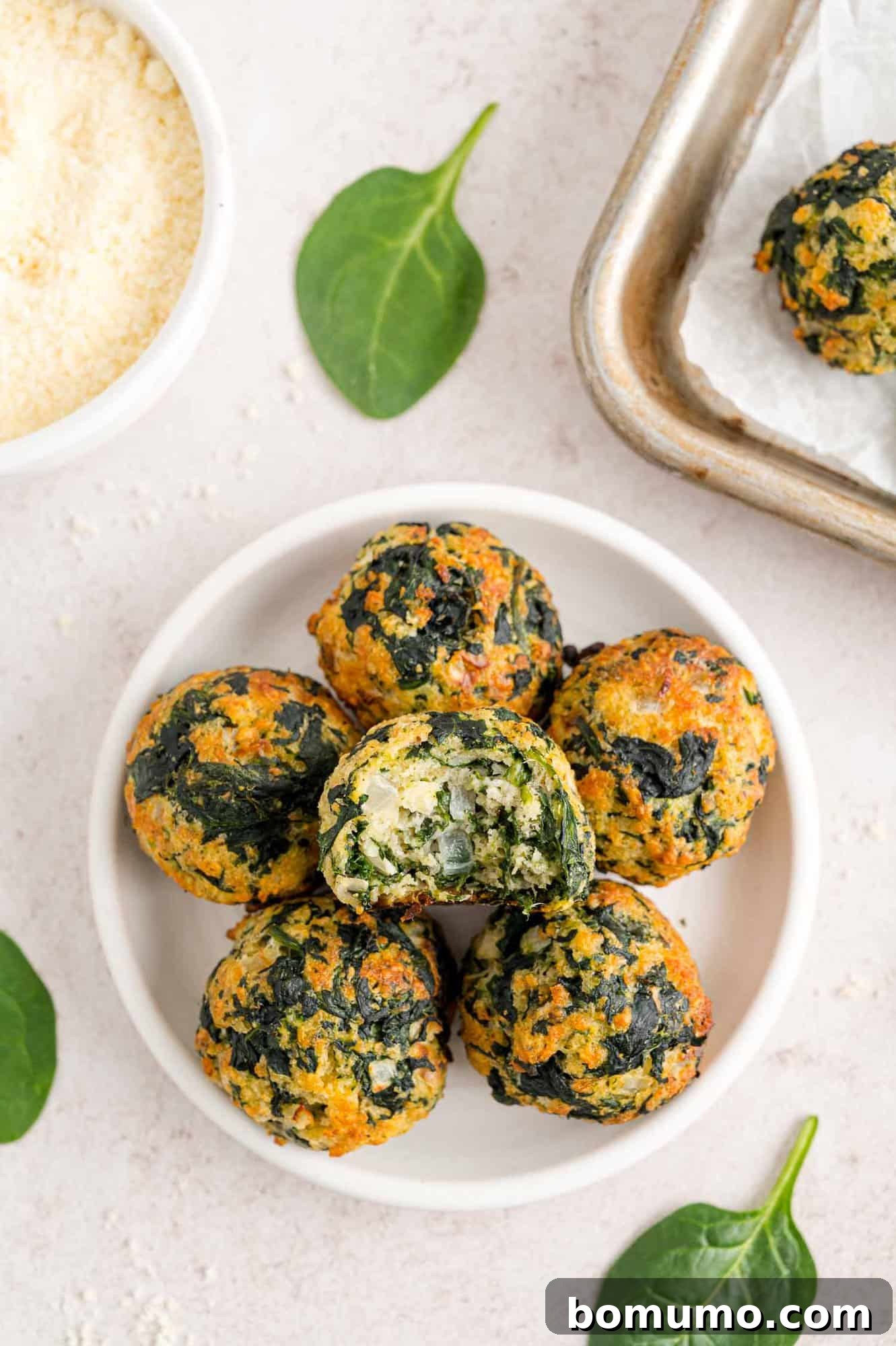 Overhead view of spinach balls stacked on a white plate, with a bite missing from the top spinach ball, next to a bowl of dipping sauce and the corner of a baking sheet.