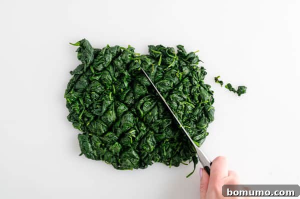 A hand uses a knife to chop cooked spinach on a white surface.