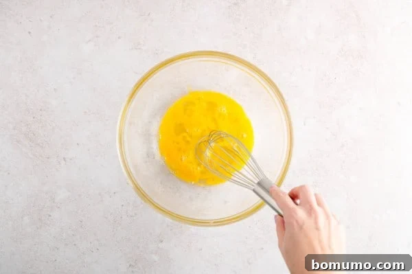 A hand uses a whisk to beat eggs in a glass bowl.