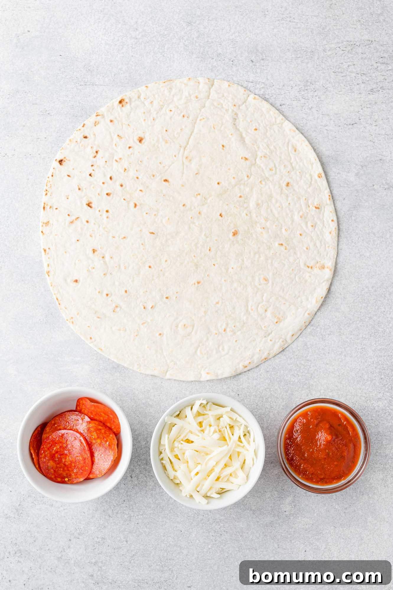 Pizza-Dilla Melt 4 A flat lay of fresh ingredients: a large flour tortilla, a bowl of pizza sauce, a mound of shredded mozzarella cheese, and a pile of pepperoni slices, ready for assembly.