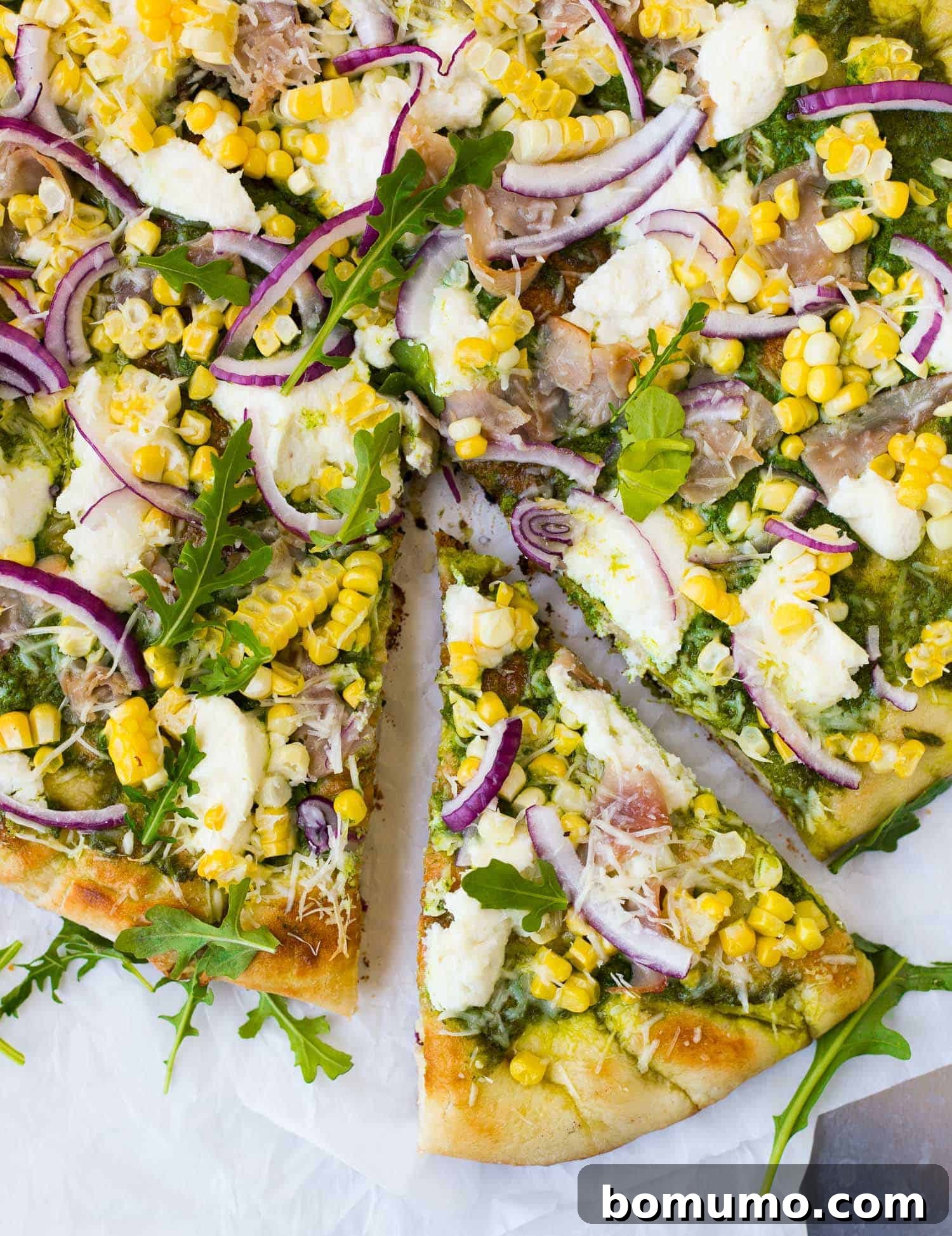 Vibrant overhead view of a grilled pizza topped generously with fresh arugula, thinly sliced red onions, savory ham, and bright sweet corn kernels. One slice has been partially removed, revealing the golden, crispy crust underneath.