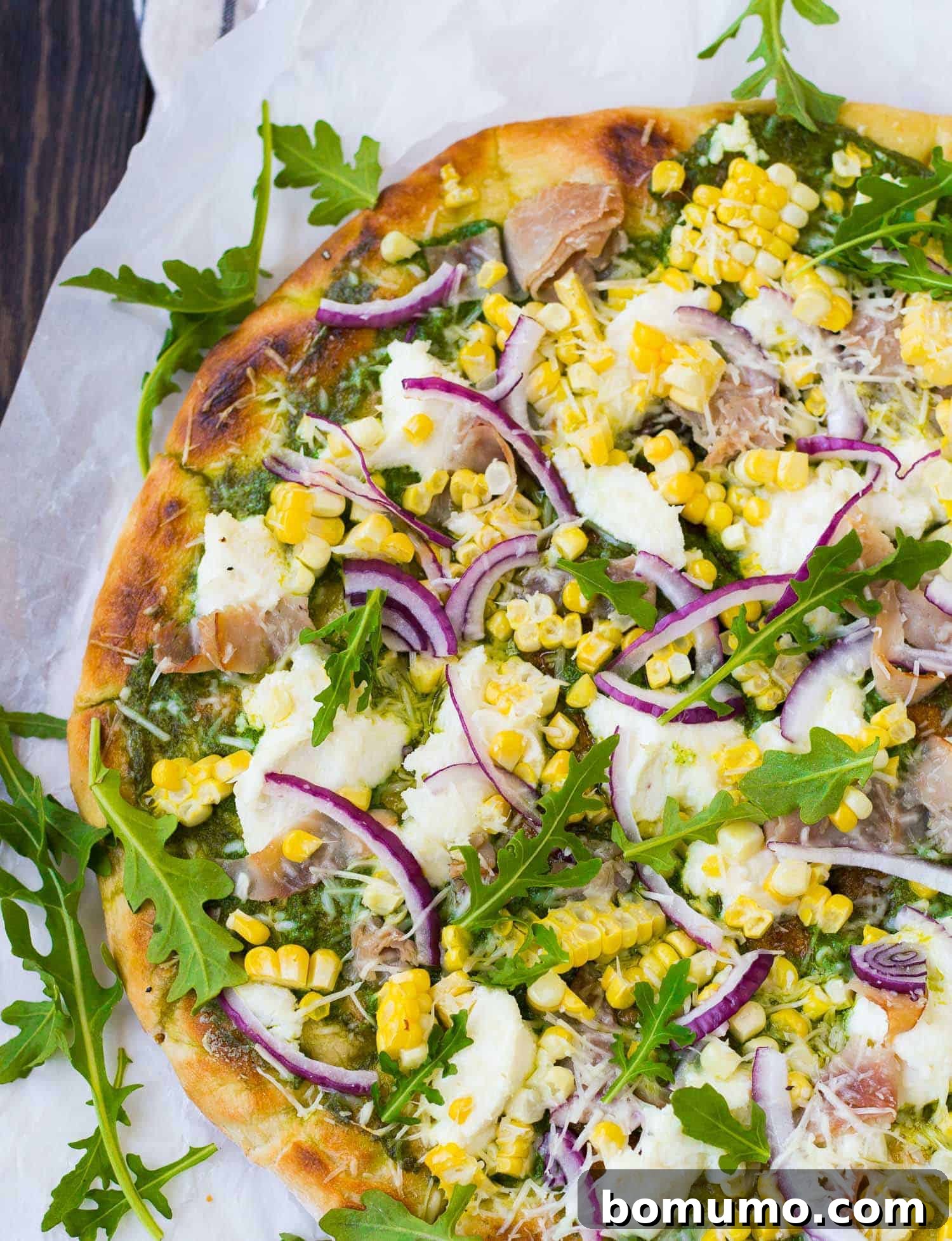 A pristine, uncooked pizza dough on parchment paper, beautifully topped with creamy ricotta cheese, vibrant arugula, thinly sliced red onion, and savory ham, awaiting its turn on the grill.
