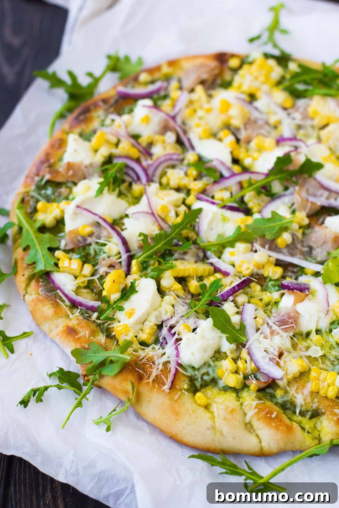 A freshly grilled pizza, served on a piece of parchment paper, adorned with vibrant arugula leaves and dollops of creamy ricotta cheese, ready to be enjoyed.