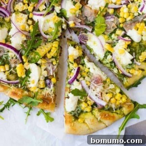 Overhead view of pizza topped with arugula, red onions, ham, and corn. One slice partially removed.