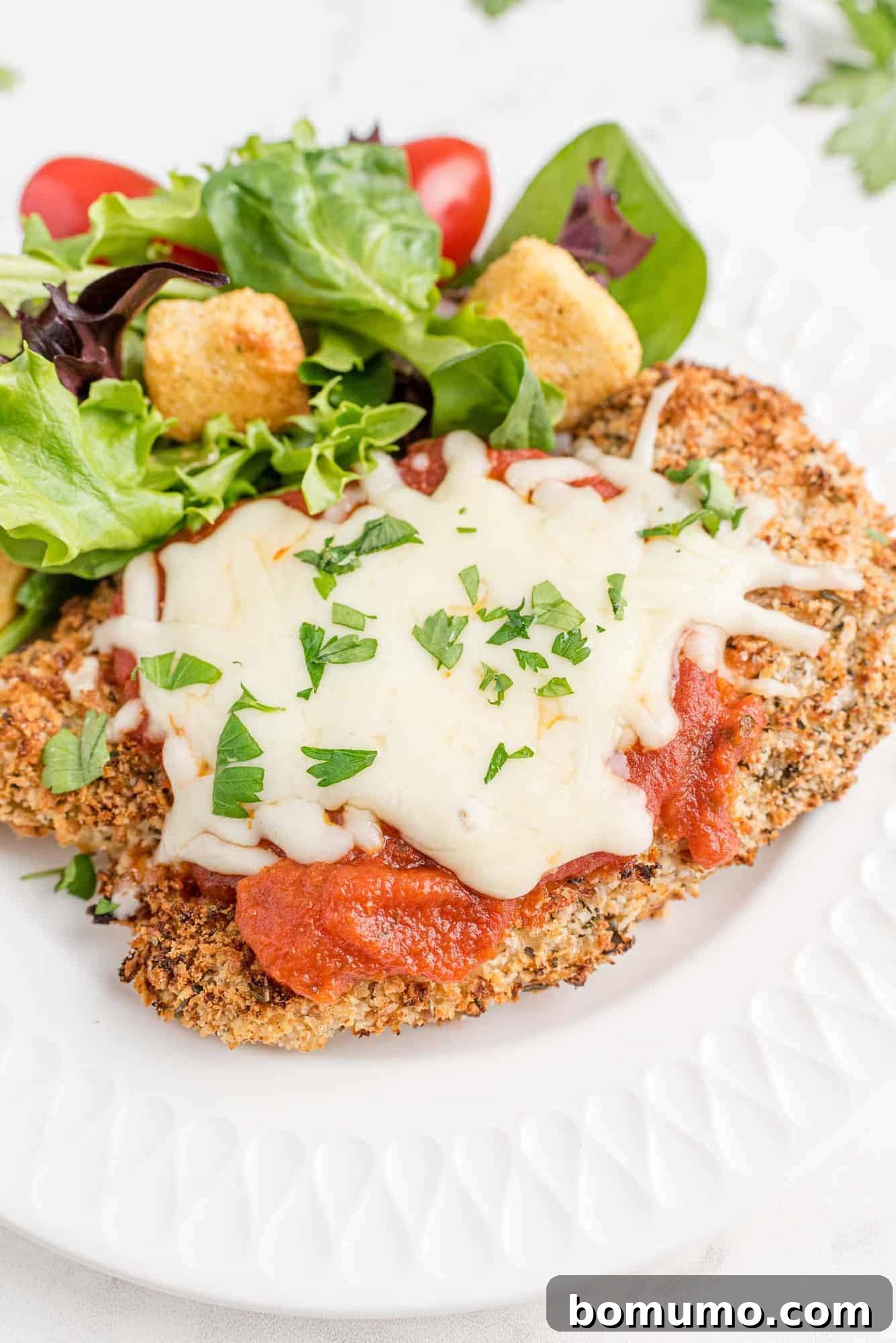 Baked chicken Parmesan on a white plate with a tossed green salad, highlighting its crispy golden crust and melted cheese.