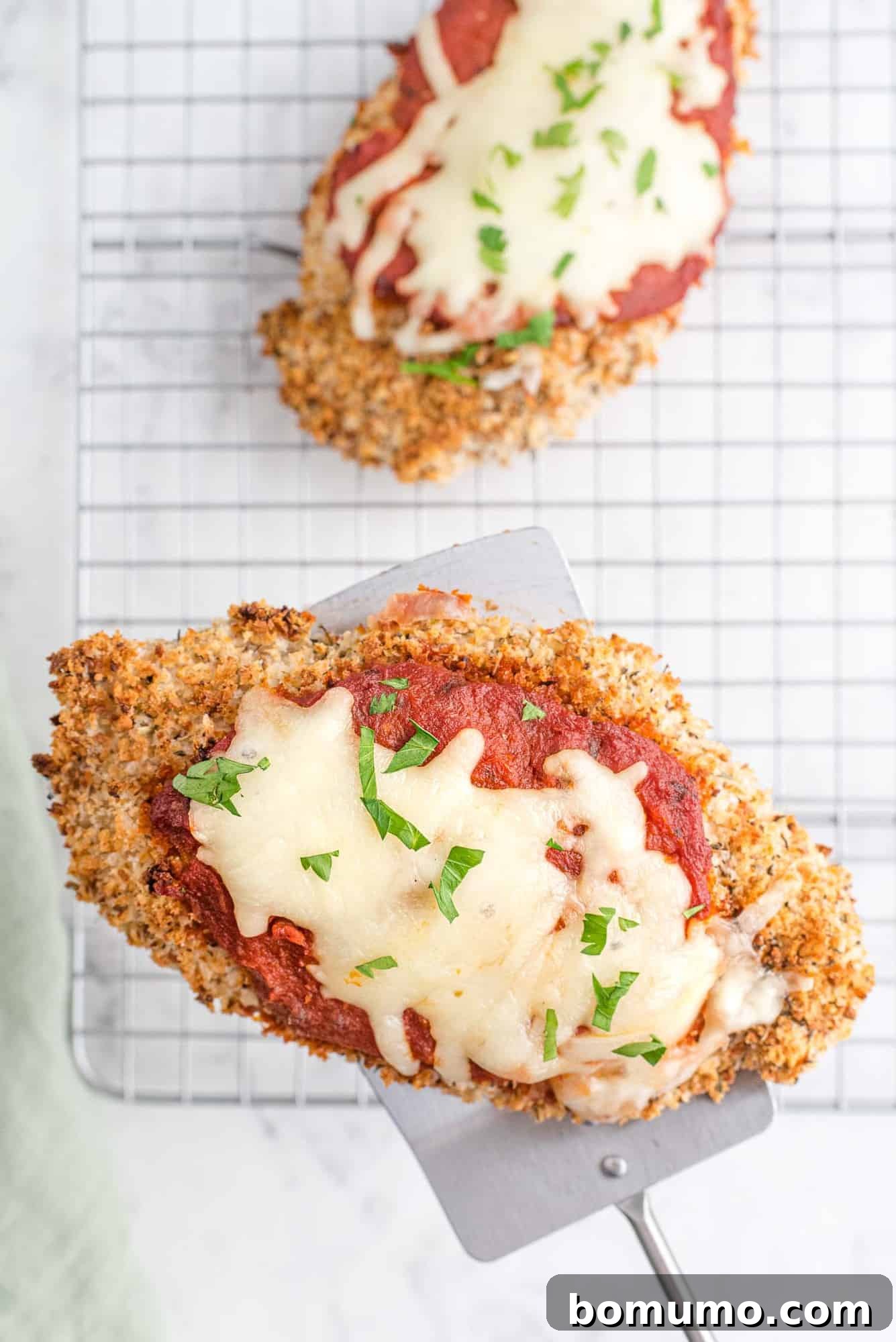 A hand using a metal spatula to carefully lift a piece of baked chicken Parmesan off a metal baking rack, ready to be served.