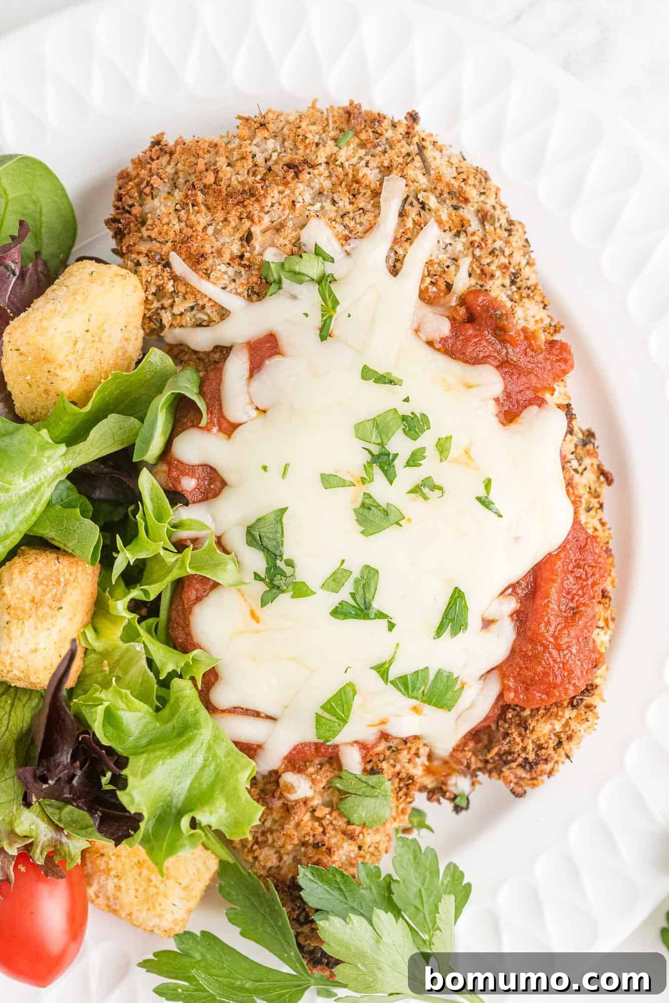 Chicken parmesan on a white plate with salad, showcasing its golden crust and delicious toppings.