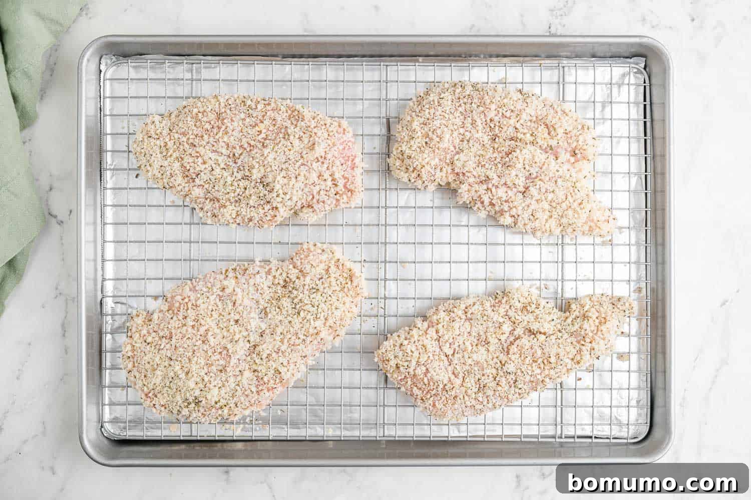 Breaded but uncooked chicken cutlets arranged on a wire rack over a baking sheet, ready for the oven.