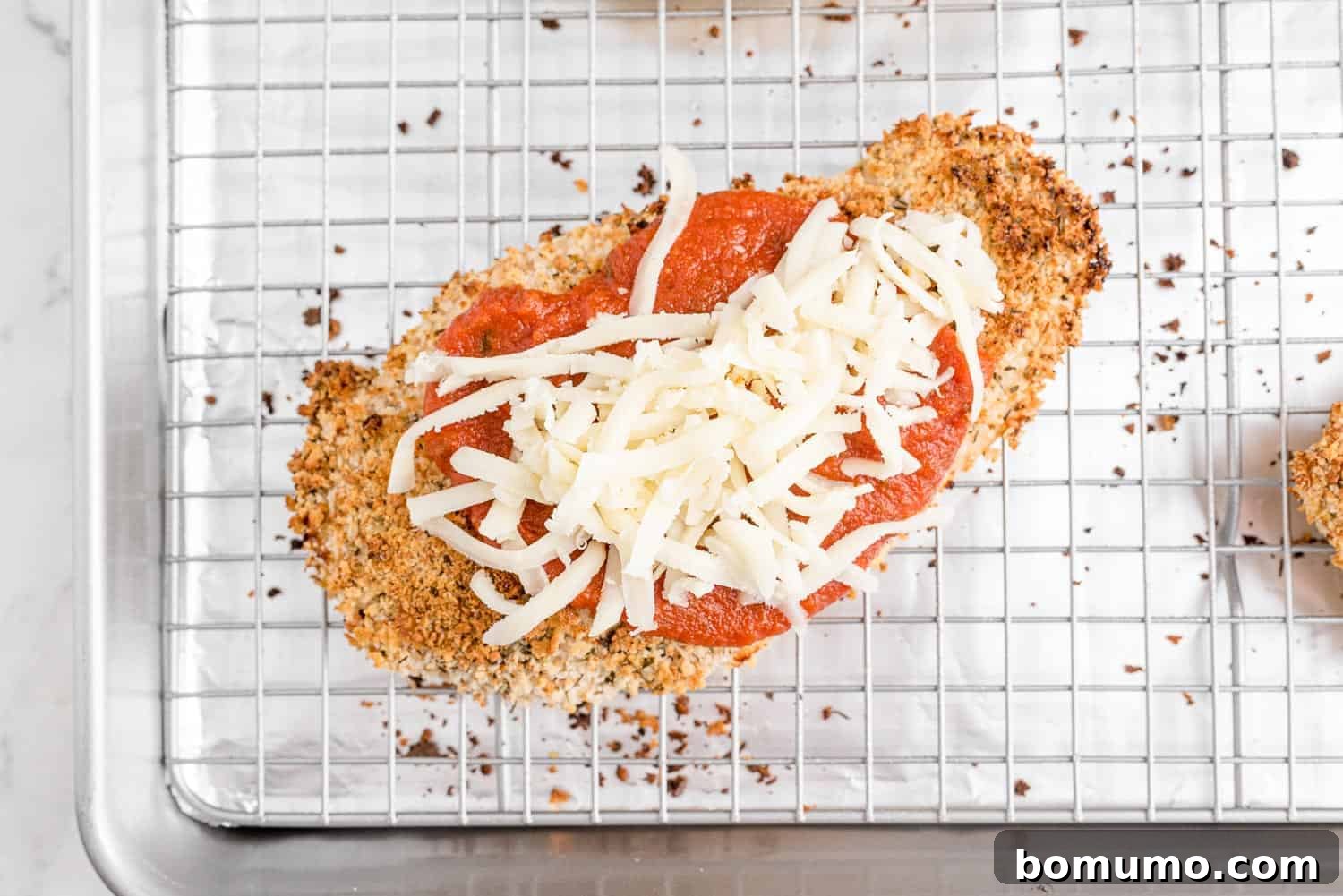 A baked chicken cutlet topped with rich marinara sauce and a generous sprinkle of shredded mozzarella cheese, ready for final melting.