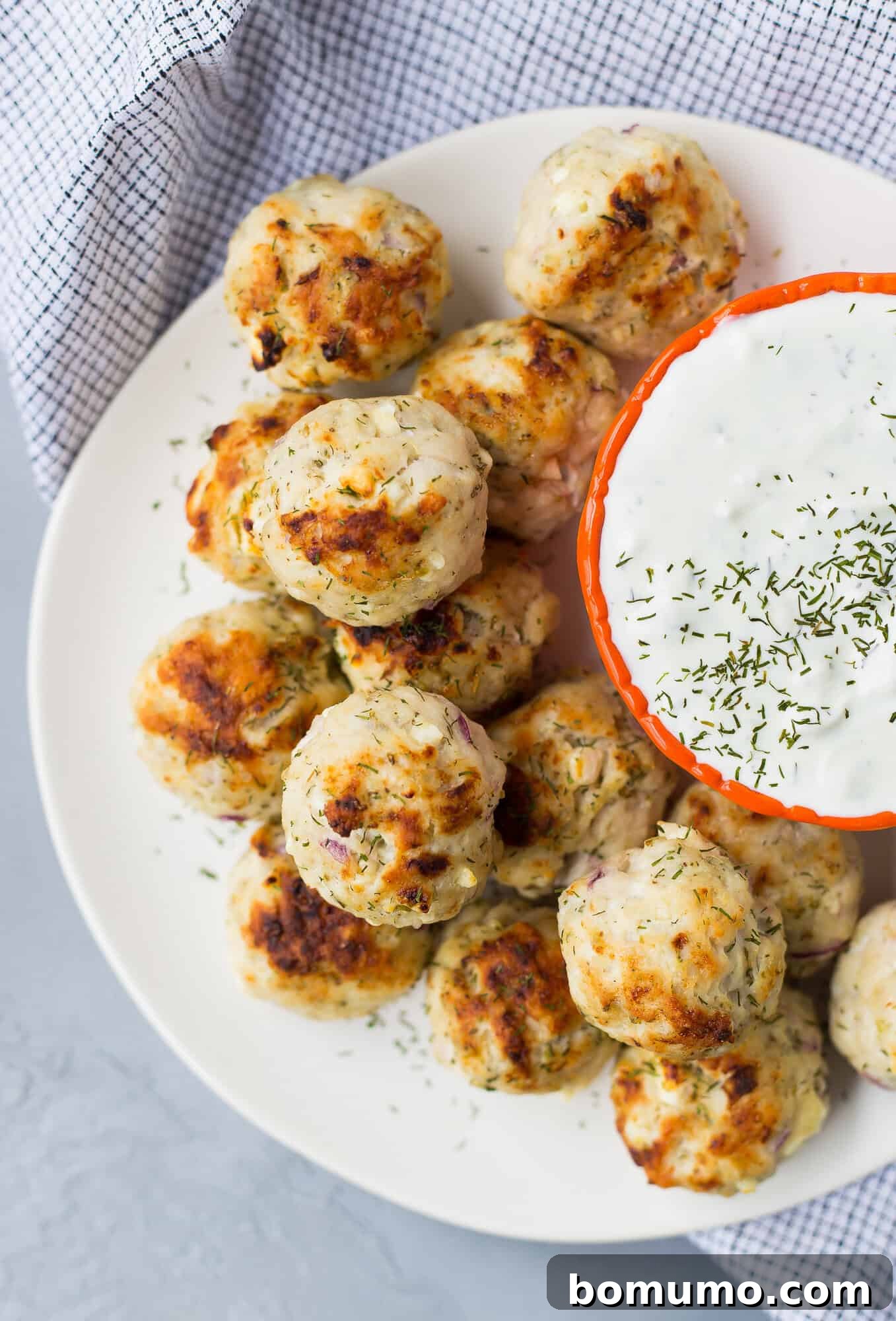 A white plate with several Greek turkey meatballs next to a small bowl of creamy dipping sauce, sprinkled with herbs, on a gray surface with a checkered cloth.
