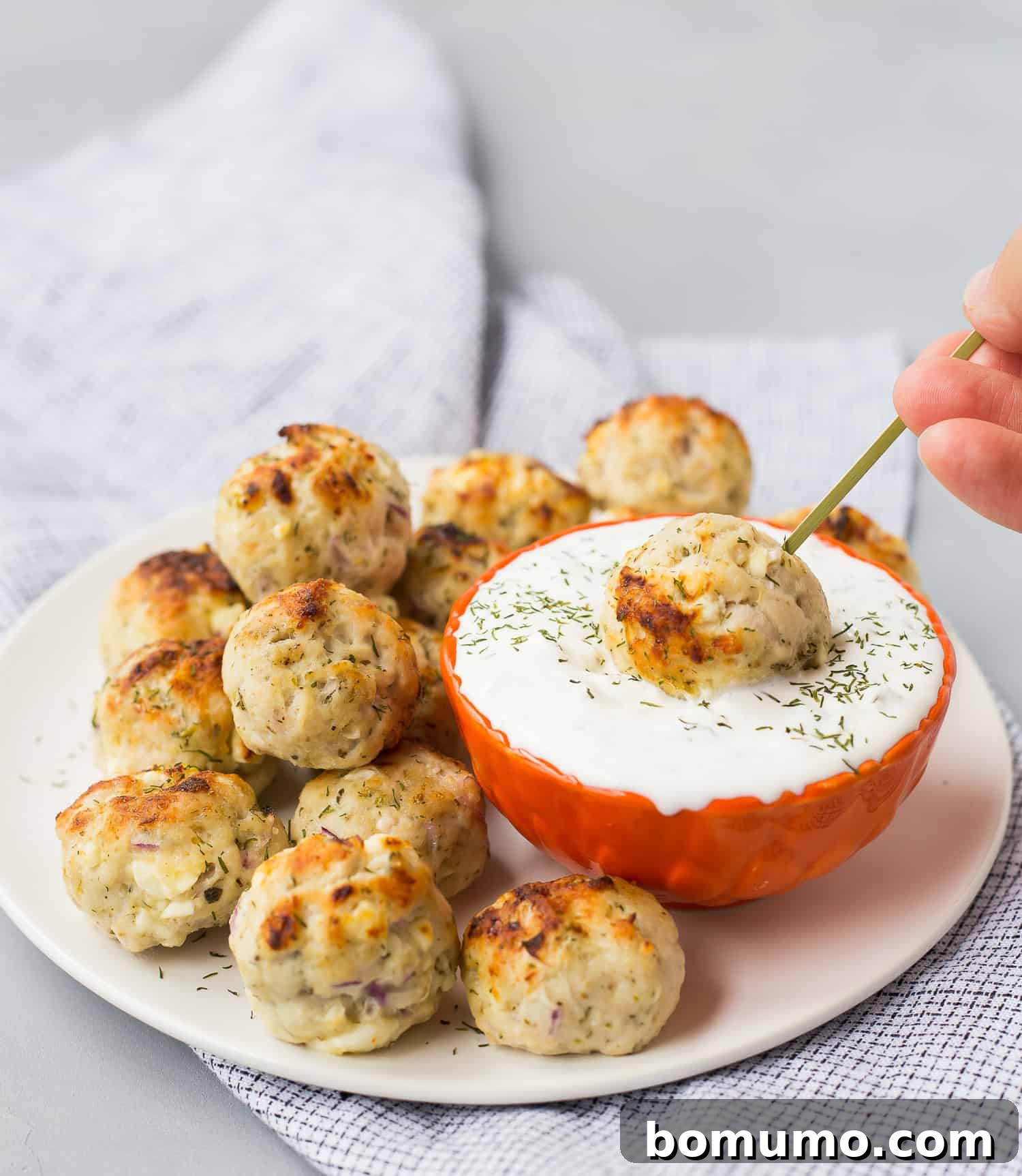 A plate of baked greek turkey meatballs is served with a bowl of white dipping sauce; one meatball is being dipped into the sauce using a skewer.