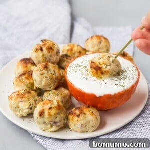 A plate of baked Greek turkey meatballs next to a bowl of white dipping sauce, with one meatball being dipped using a skewer.