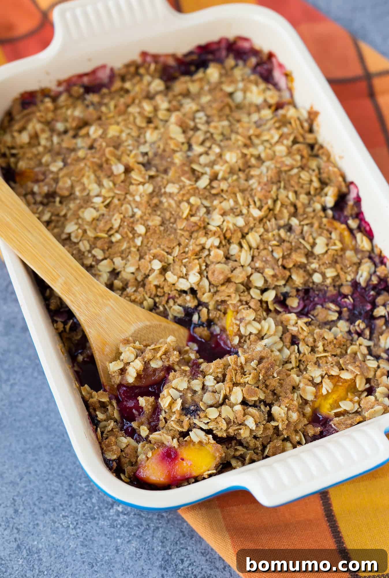 A golden-brown Peach Blueberry Crisp in a baking dish, with a large wooden spoon ready for serving. The crisp topping is perfectly baked, showcasing hints of juicy fruit peeking through.