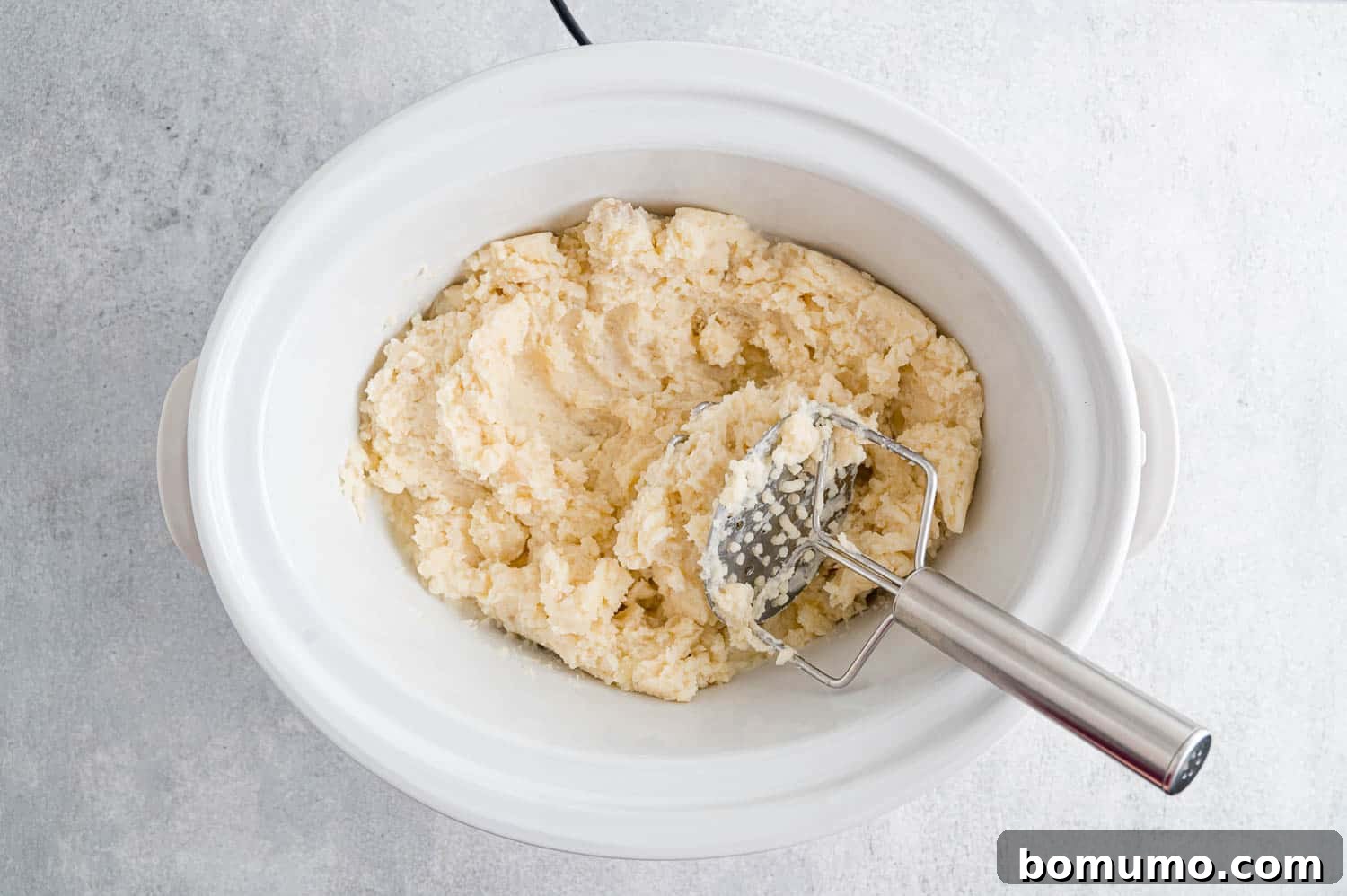 Set-It-and-Forget-It Mashed Potatoes 10 A potato masher actively mashing cooked potatoes with butter and sour cream directly in the slow cooker.