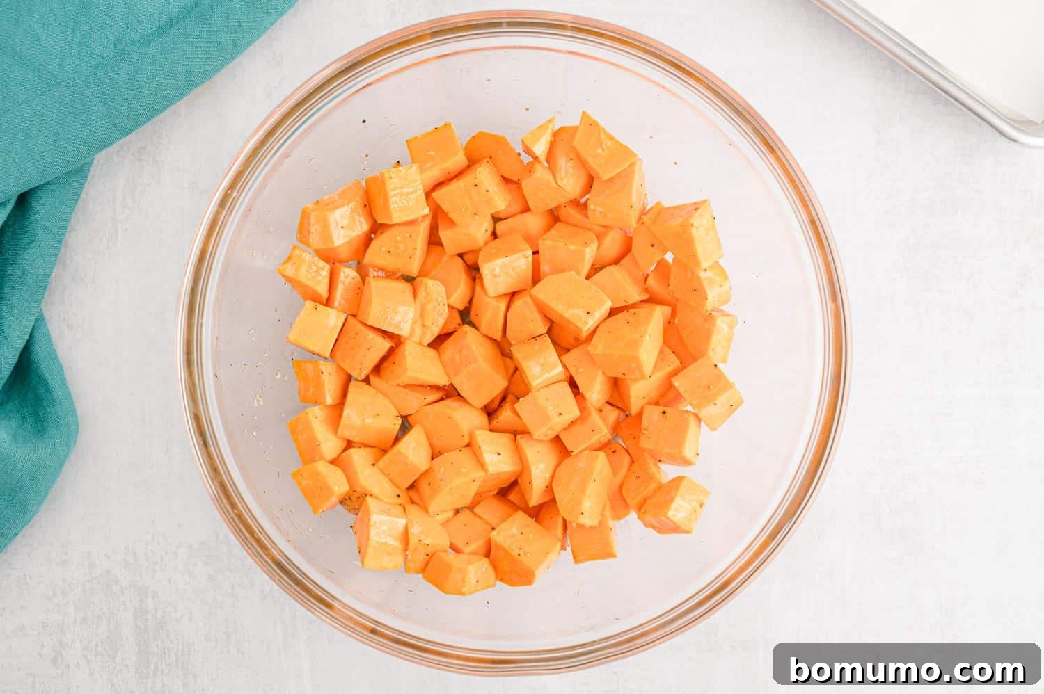 Sweet potatoes in a clear glass bowl, coated with oil and seasoning, ready for the baking sheet.