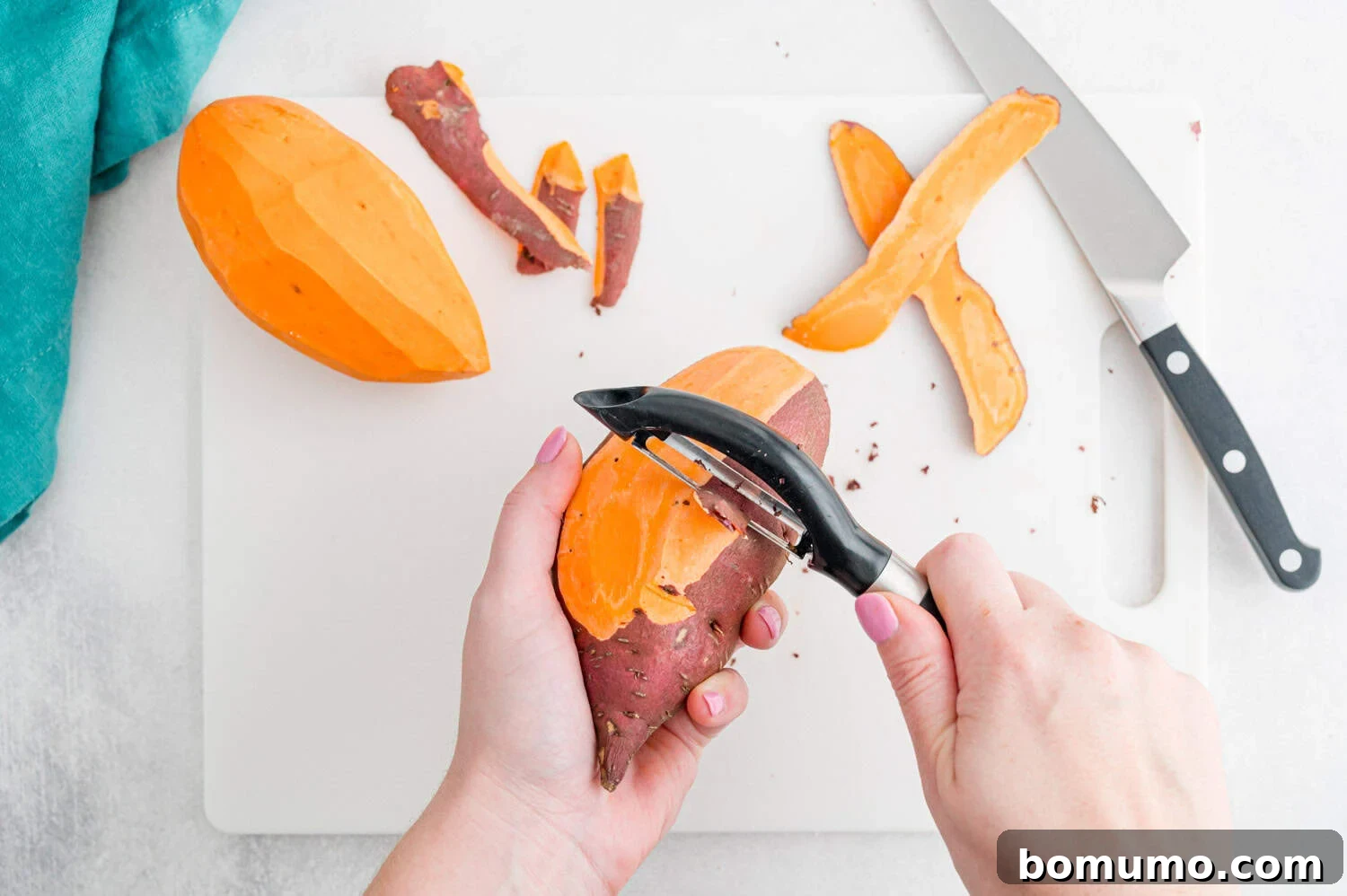 A person peels a sweet potato with a vegetable peeler on a white cutting board next to a knife, peels, and a partially peeled sweet potato.