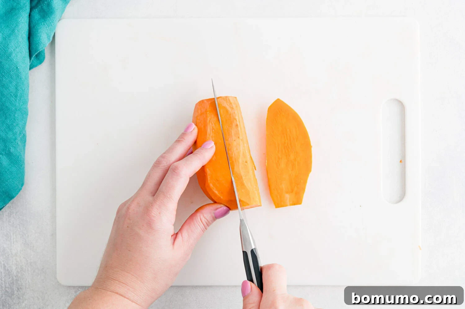 A person slices a piece of peeled sweet potato on a white cutting board with a kitchen knife.
