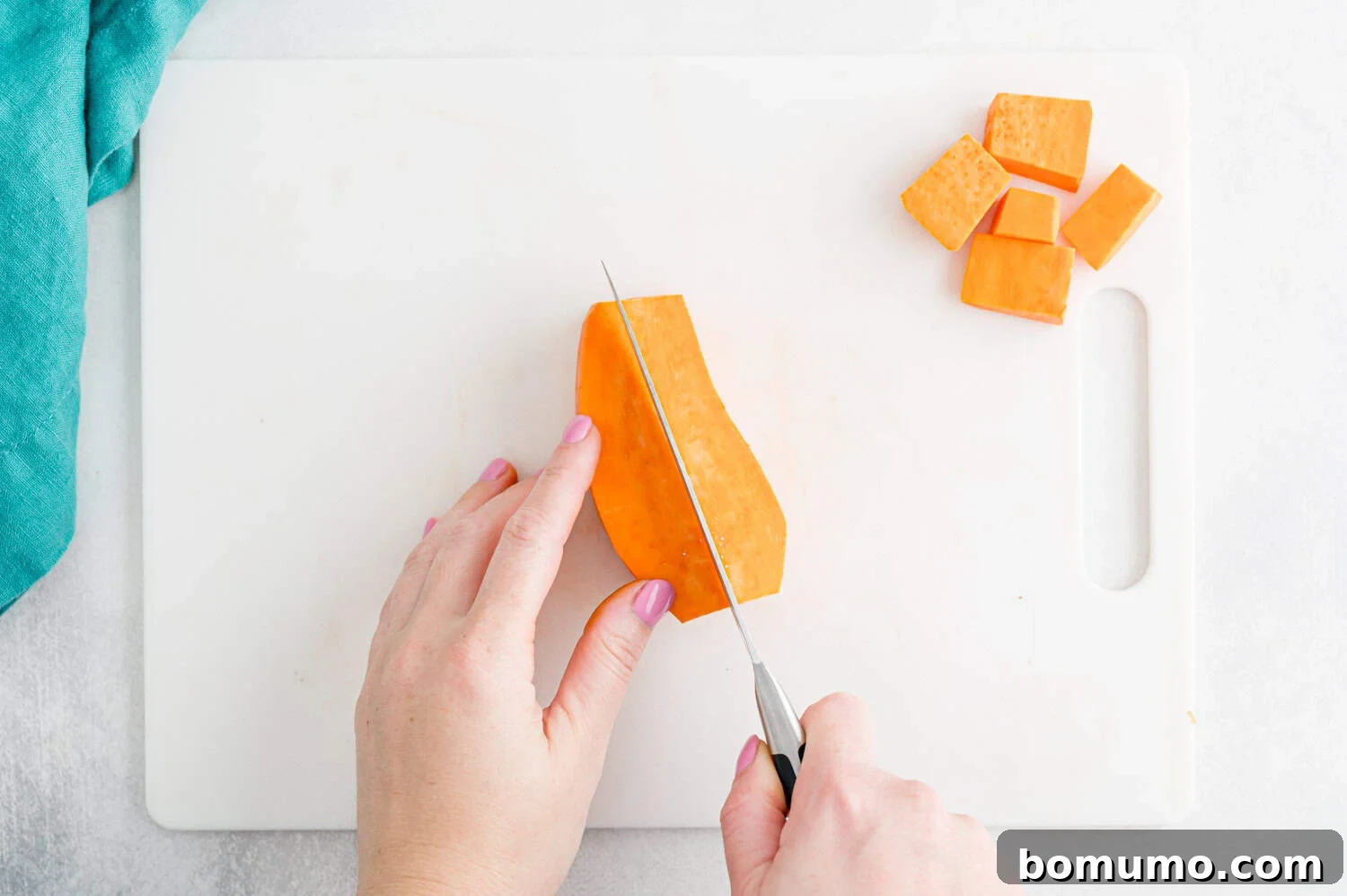 A person slices a peeled sweet potato on a white cutting board, with several cubed pieces set aside; a blue cloth is visible in the corner, illustrating the dicing process.