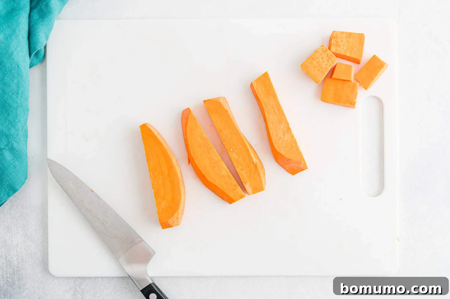 A chef’s knife and sliced sweet potato wedges and cubes on a white cutting board with a teal cloth nearby, ready for seasoning.