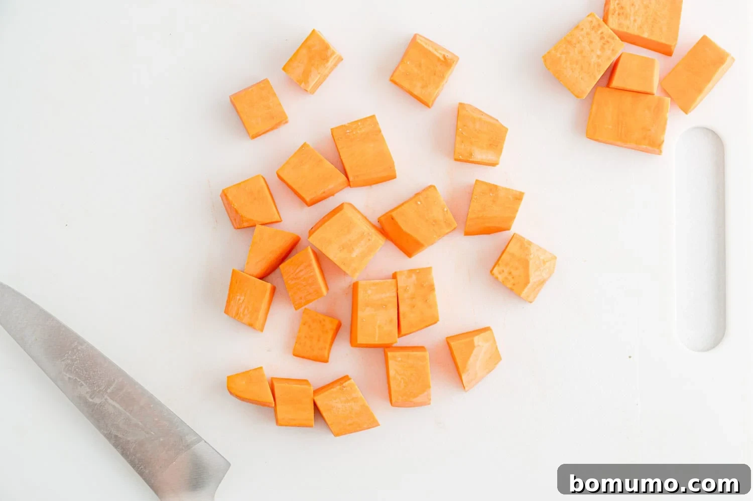 Cubed pieces of sweet potato on a white cutting board next to a kitchen knife, demonstrating the final dice size.