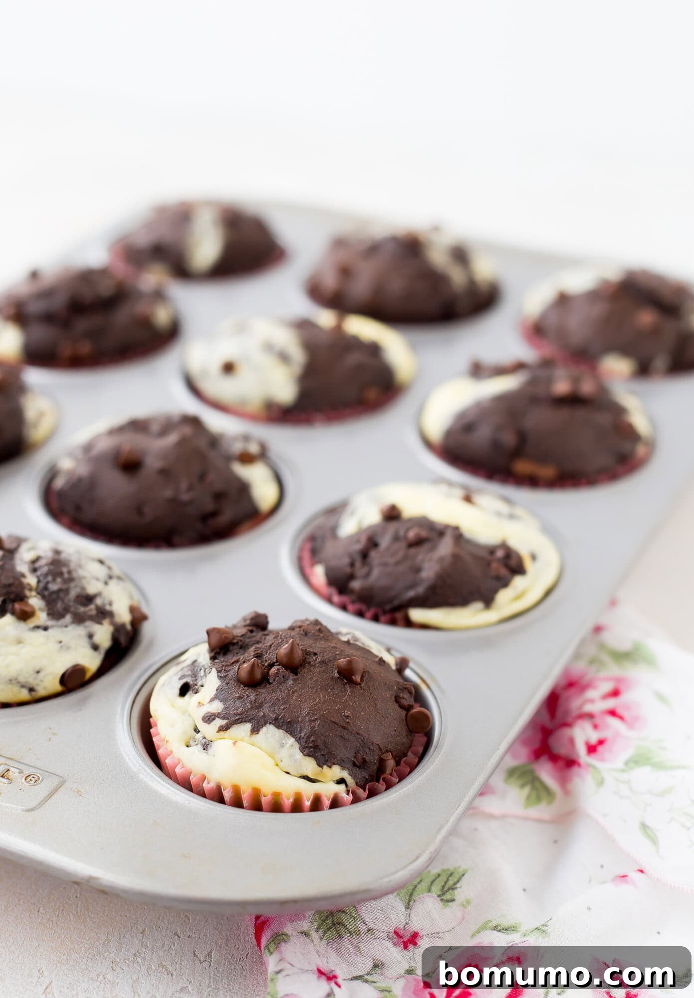 A muffin tin filled with freshly baked marble chocolate chip cream cheese muffins sits on a floral cloth.