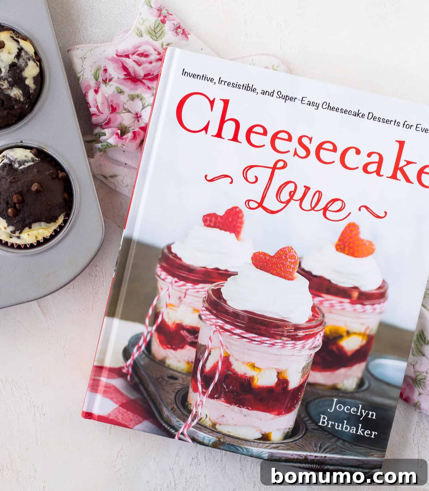 A cookbook titled Cheesecake Love by Jocelyn Brubaker is on a floral cloth next to a muffin tin with two baked muffins.