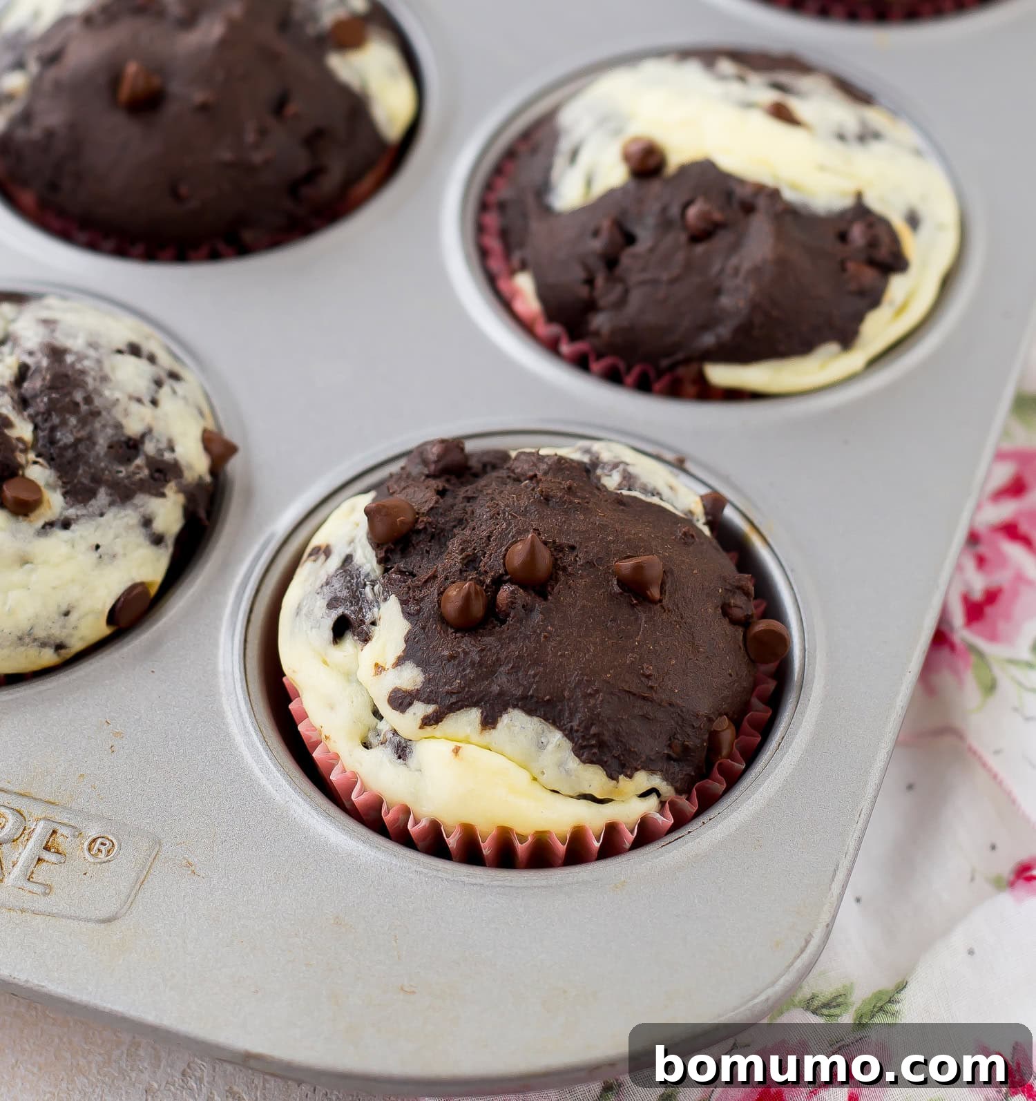 A muffin tin with marbled chocolate and cream cheese muffins, each topped with chocolate chips, on a floral-patterned cloth.
