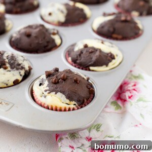 A muffin tin filled with marbled chocolate cream cheese muffins, some topped with chocolate chips, rests on a floral-patterned cloth.