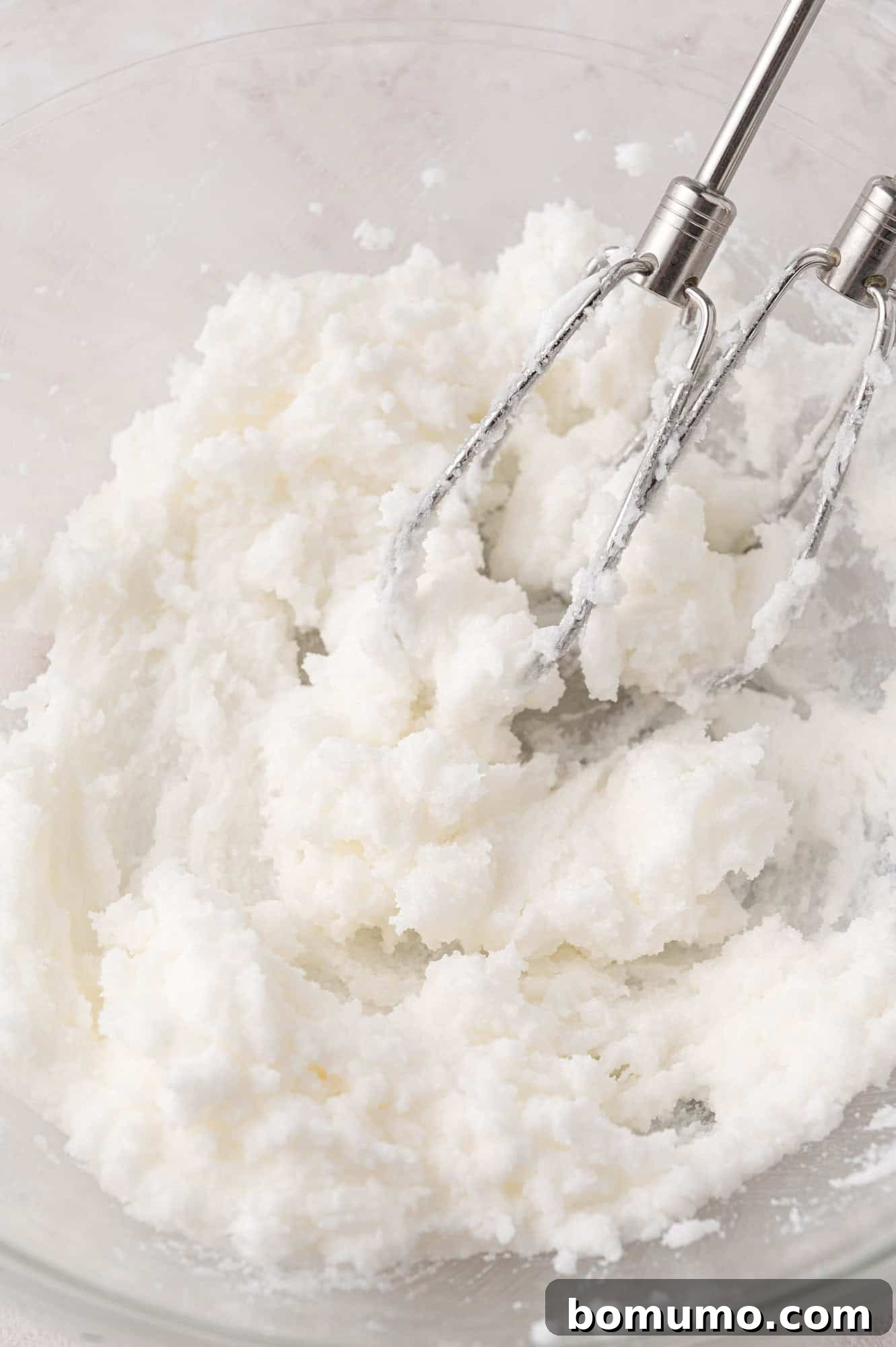 A close-up of two metal beaters mixing white, fluffy shortening in a glass bowl, demonstrating the creaming process.
