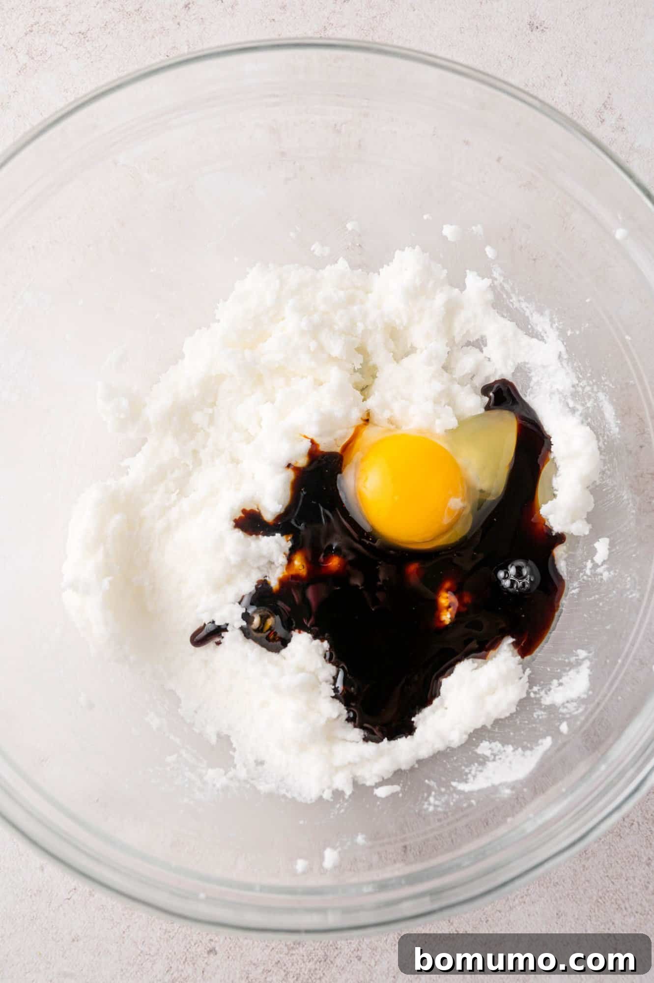 A glass bowl with creamed shortening and sugar, alongside a cracked egg and dark molasses, ready to be mixed into the cookie dough.