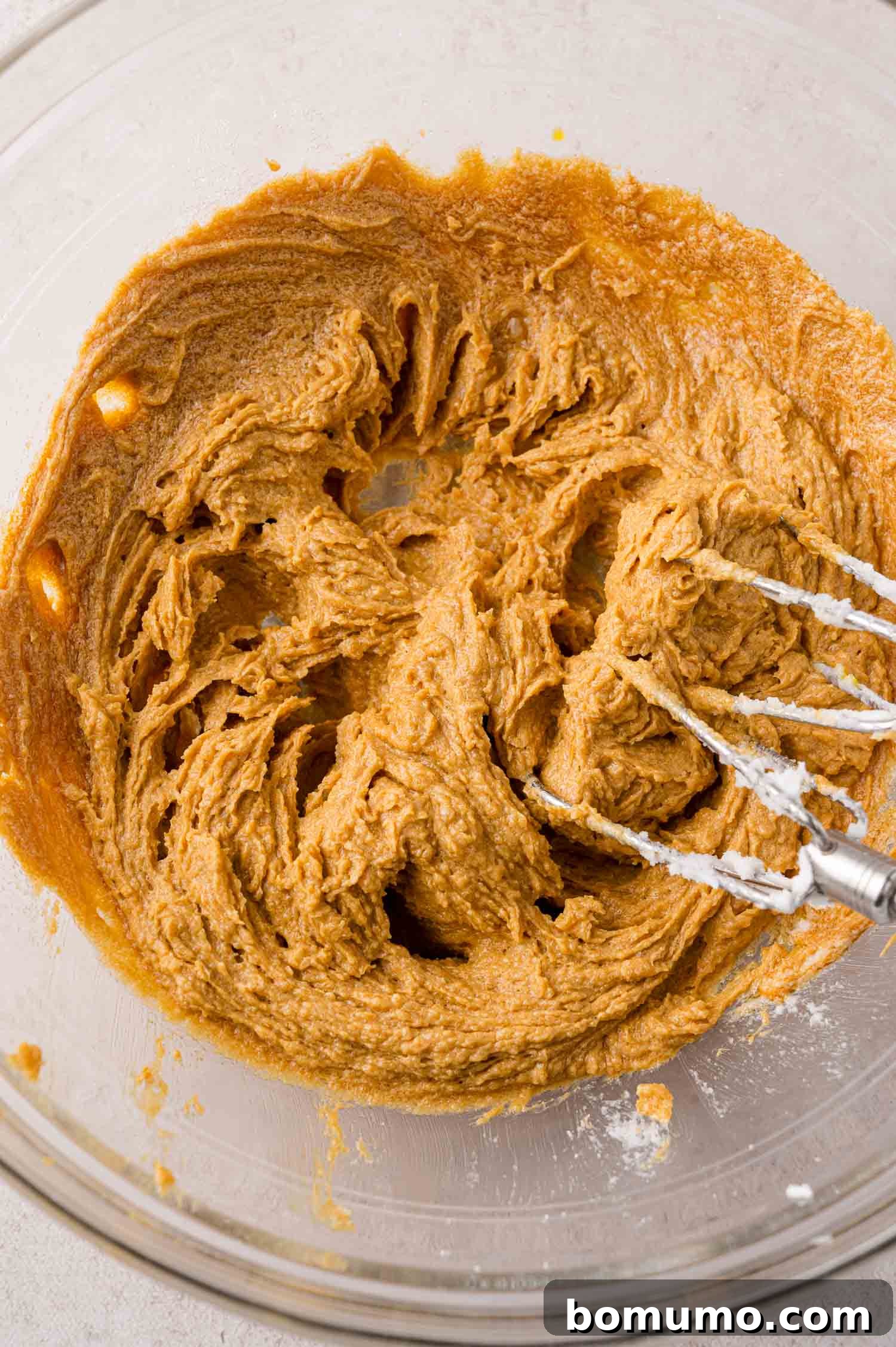 A glass bowl containing cookie dough being mixed with a hand mixer, showing the texture of the developing dough.