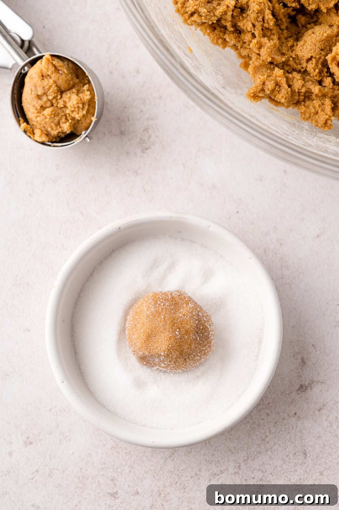 A ball of cookie dough is gently placed in a white bowl of granulated sugar, ready to be rolled and coated for baking.